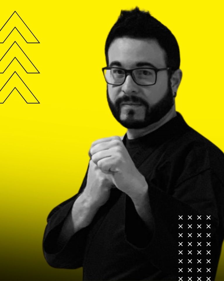 Michael Dietl Porträt Ein Mann mit Brille in schwarzer Kleidung vor einem gelben Hintergrund zeigt eine Kampfkunstpose.